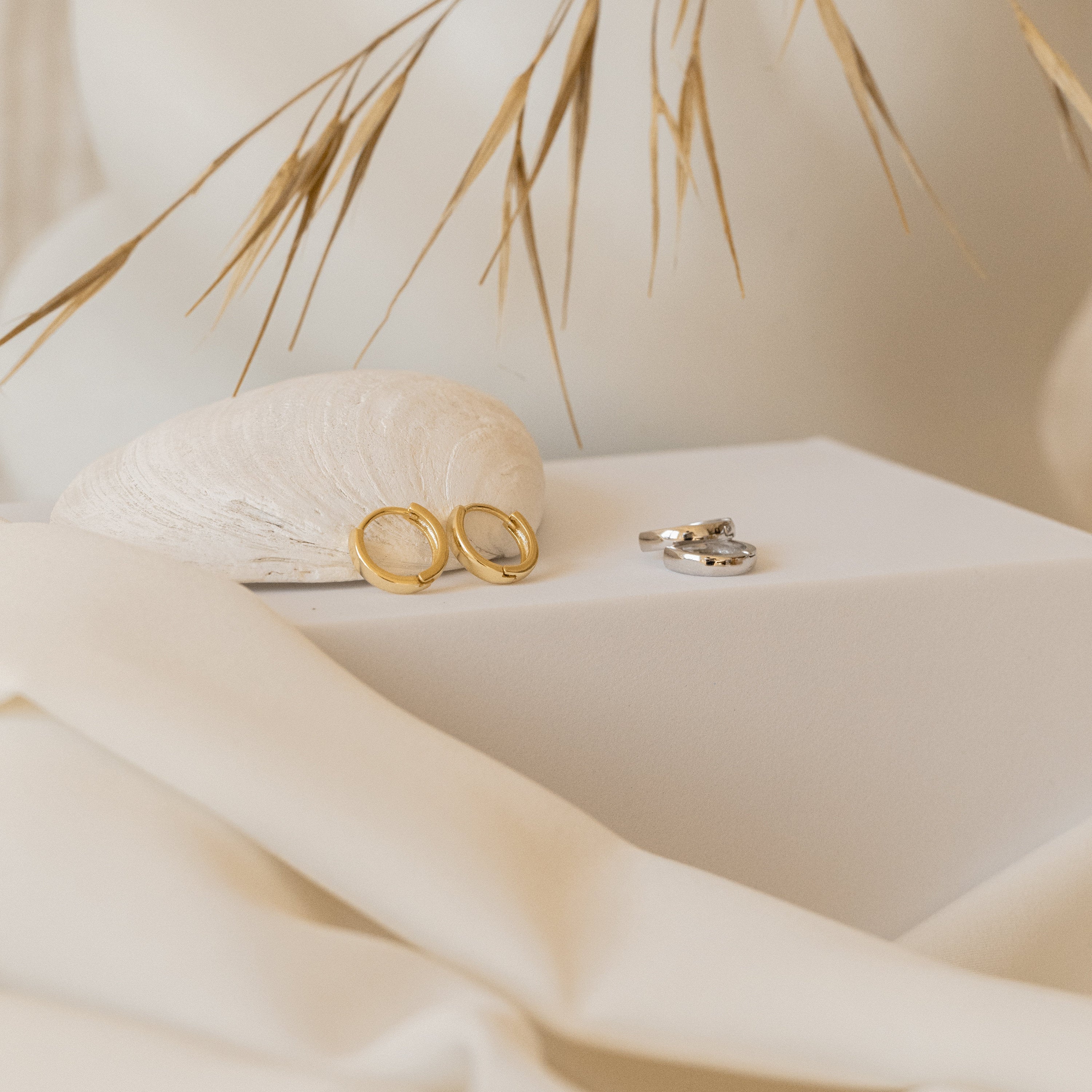 Sterling silver Editor II Huggies displayed on a white surface with a seashell and dried grass in the background, alongside classic hoop earrings perfect for everyday wear.