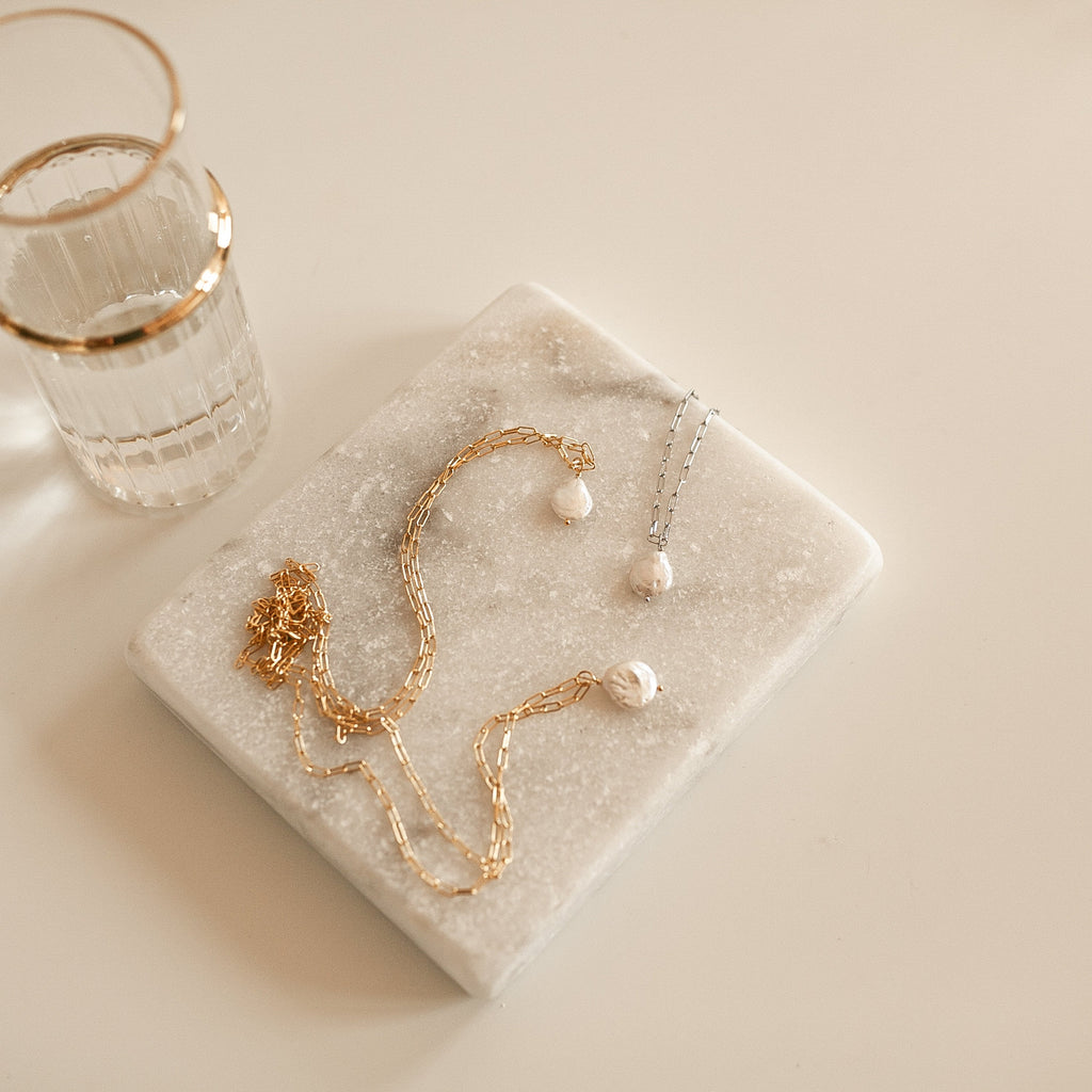 Gold and silver pearl link chain necklaces on a marble surface with a glass of water.
