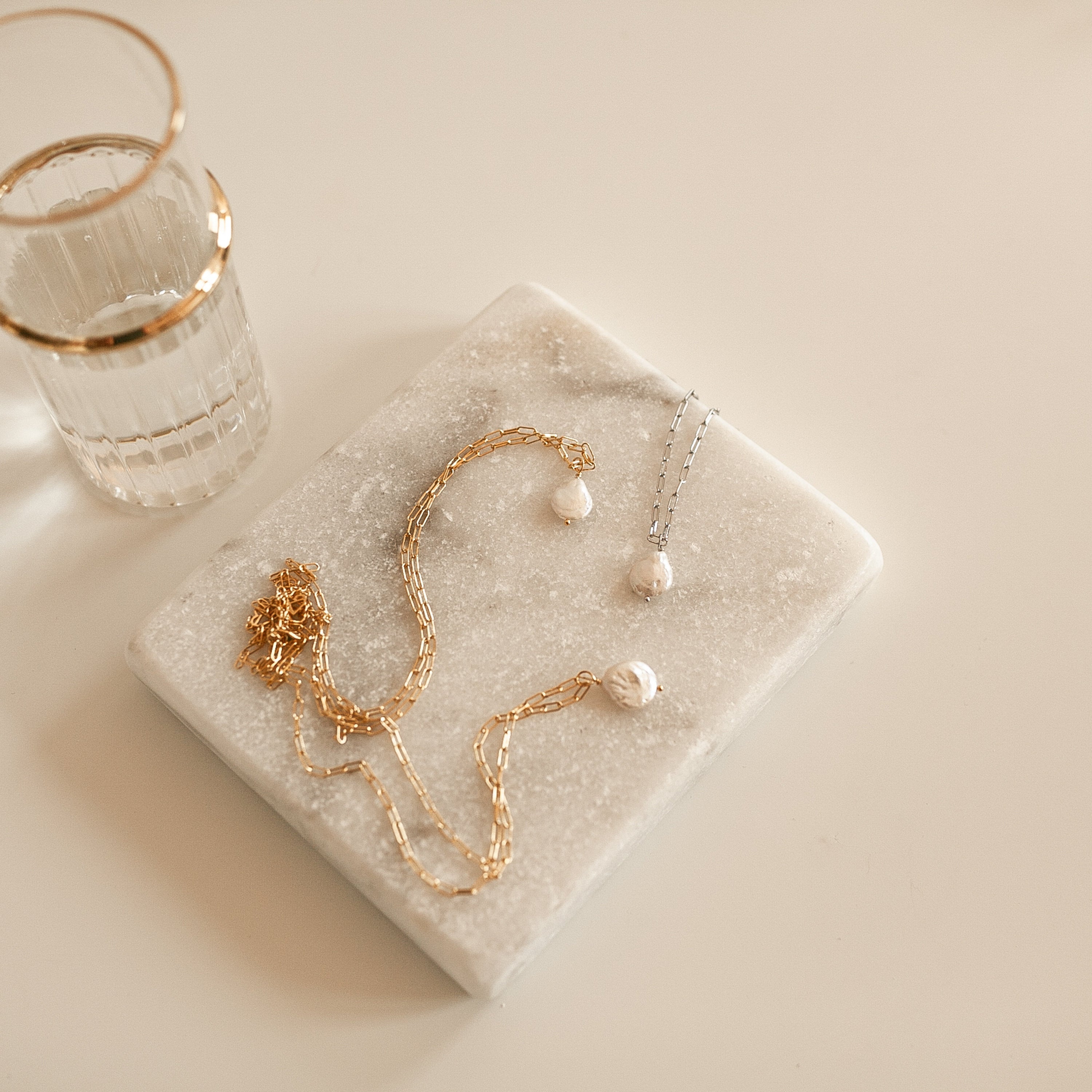 Gold and silver pearl link chain necklaces on a marble surface with a glass of water.