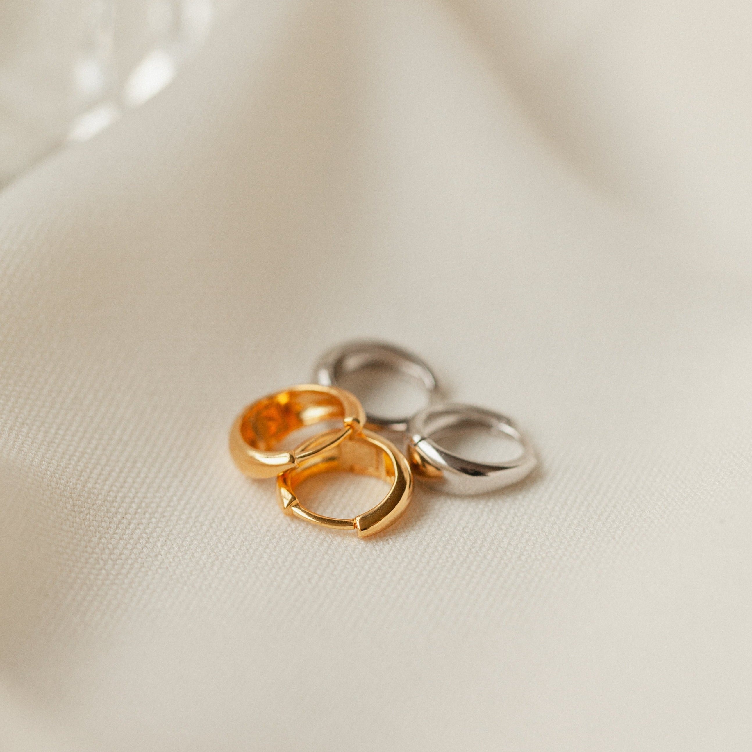 Two gold and two silver Lucy Hinged Huggies rest on a soft, light beige fabric background.