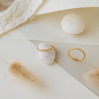 The Agate Diamond Rings Set, featuring two elegant gold rings, is displayed on white stones with soft neutral textures and dried grass in the background.