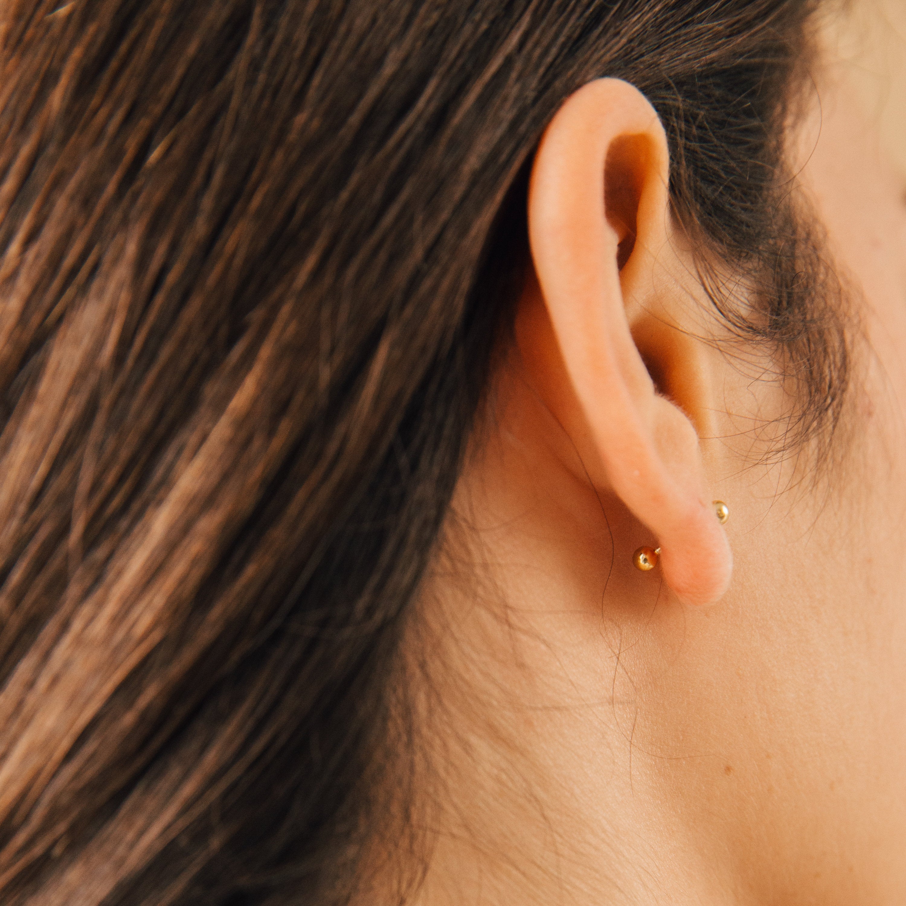 Close-up of a person’s ear with brown hair wearing the Nora Duo Studs—two small gold ball stud earrings—perfect as classic everyday earrings.