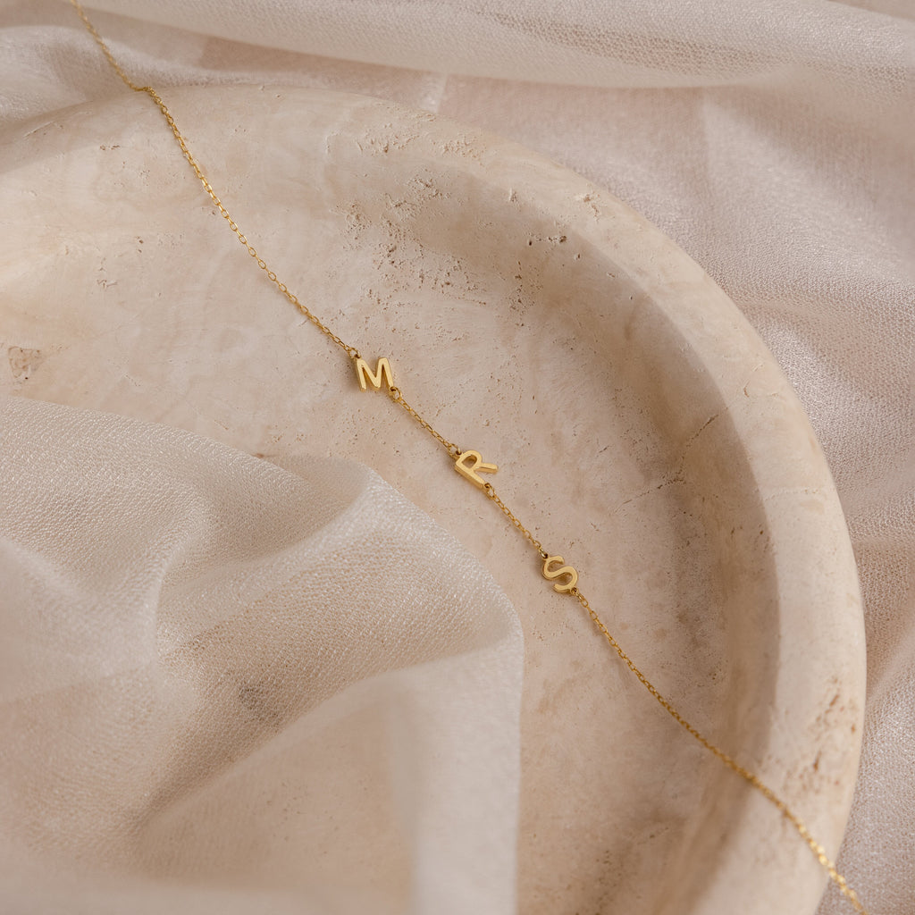 Sideways Initial Necklace with M, R, and S letter charms shown on a marble tray with sheer fabric—a unique piece that's perfect for Christmas gifting.