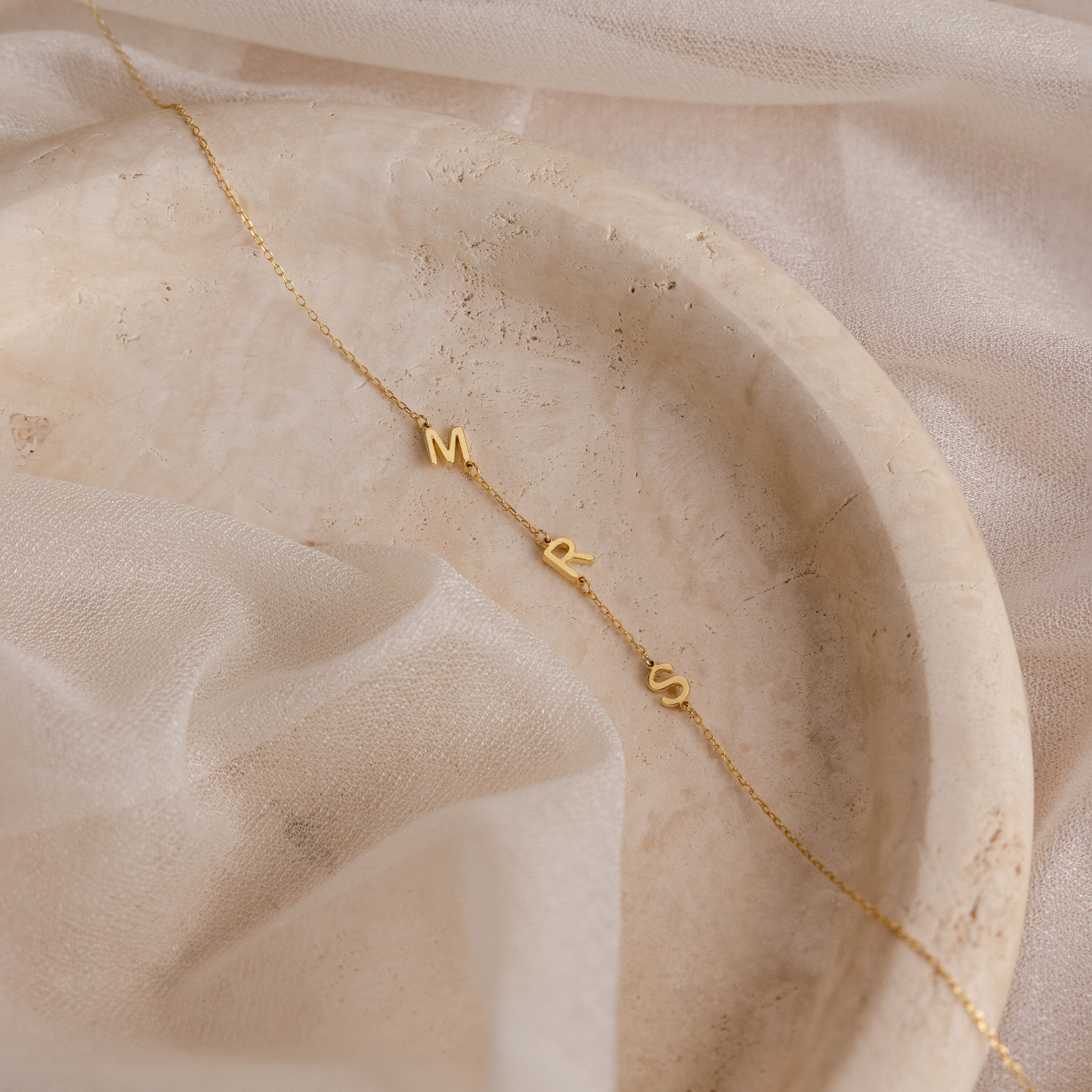 Sideways Initial Necklace with M, R, and S letter charms shown on a marble tray with sheer fabric—a unique piece that's perfect for Christmas gifting.