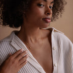 A woman with a neutral expression touches her chest, wearing a cream ribbed top and the Venice Sideways Initial Necklace.