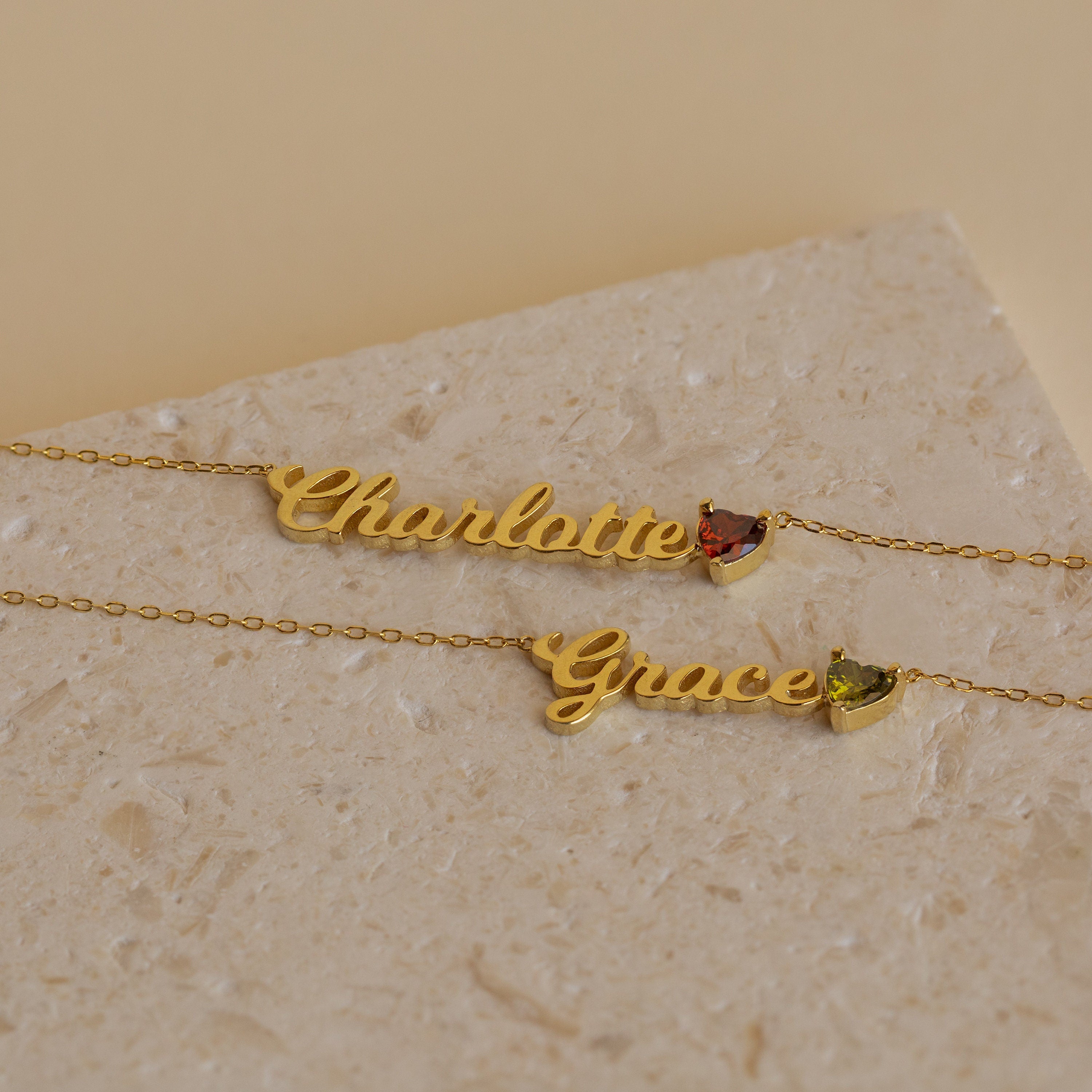 Two gold Mellow Name Heart Birthstone Necklaces, one with "Charlotte" and a red gem, the other with "Grace" and a green gem, displayed on beige stone—an elegant showcase of minimalist jewelry.