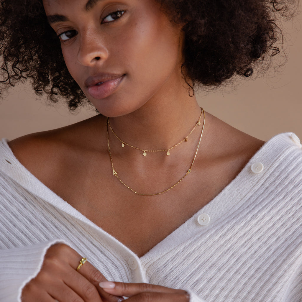 A woman in a white sweater wears layered gold necklaces, featuring the Birthstone Initial Necklace in Box Chain and a gold ring, as she looks at the camera.