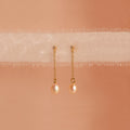 Isha Pink Pearl Earrings feature gold chains with pearl drops, elegantly displayed on soft, sheer fabric against a peach background—showcasing the timeless charm of these delicate dangling earrings.