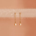 Isha Pink Pearl Earrings feature gold chains with pearl drops, elegantly displayed on soft, sheer fabric against a peach background—showcasing the timeless charm of these delicate dangling earrings.