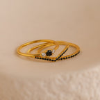 The Black Curve Ring Set features three elegant gold rings—a sparkling eternity band and two bold black diamond rings—stacked together on a subtly textured surface.