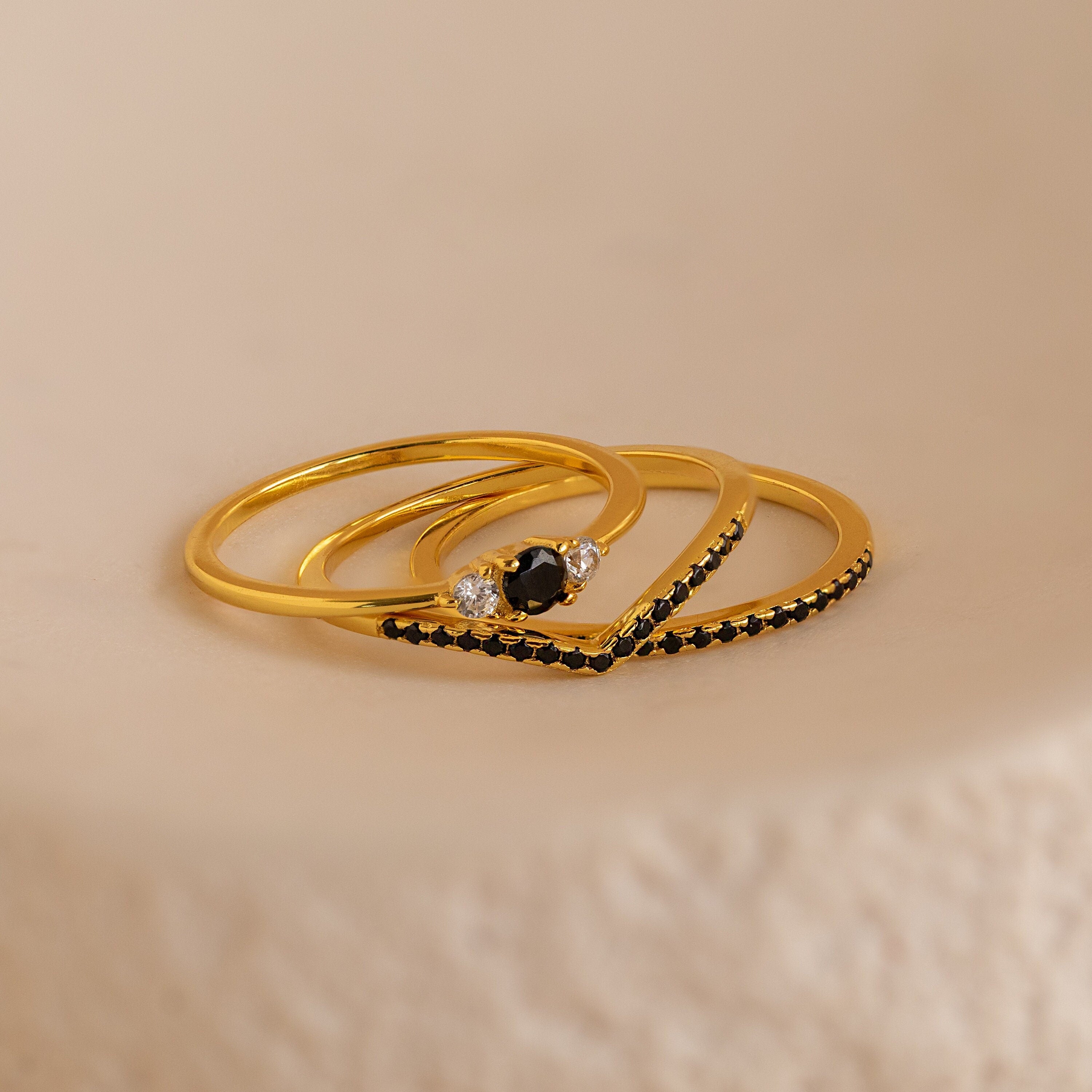 The Black Curve Ring Set features three elegant gold rings—a sparkling eternity band and two bold black diamond rings—stacked together on a subtly textured surface.