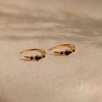 The Amaya Black Marquise Threaders feature gold hoops with black stone accents, elegantly displayed on a beige textured surface—an exquisite addition to any jewelry collection.