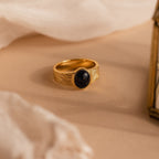 The Celestial Black Signet Ring features a midnight blue sandstone oval stone and gold finish, displayed on soft beige fabric—a stunning statement jewelry piece.