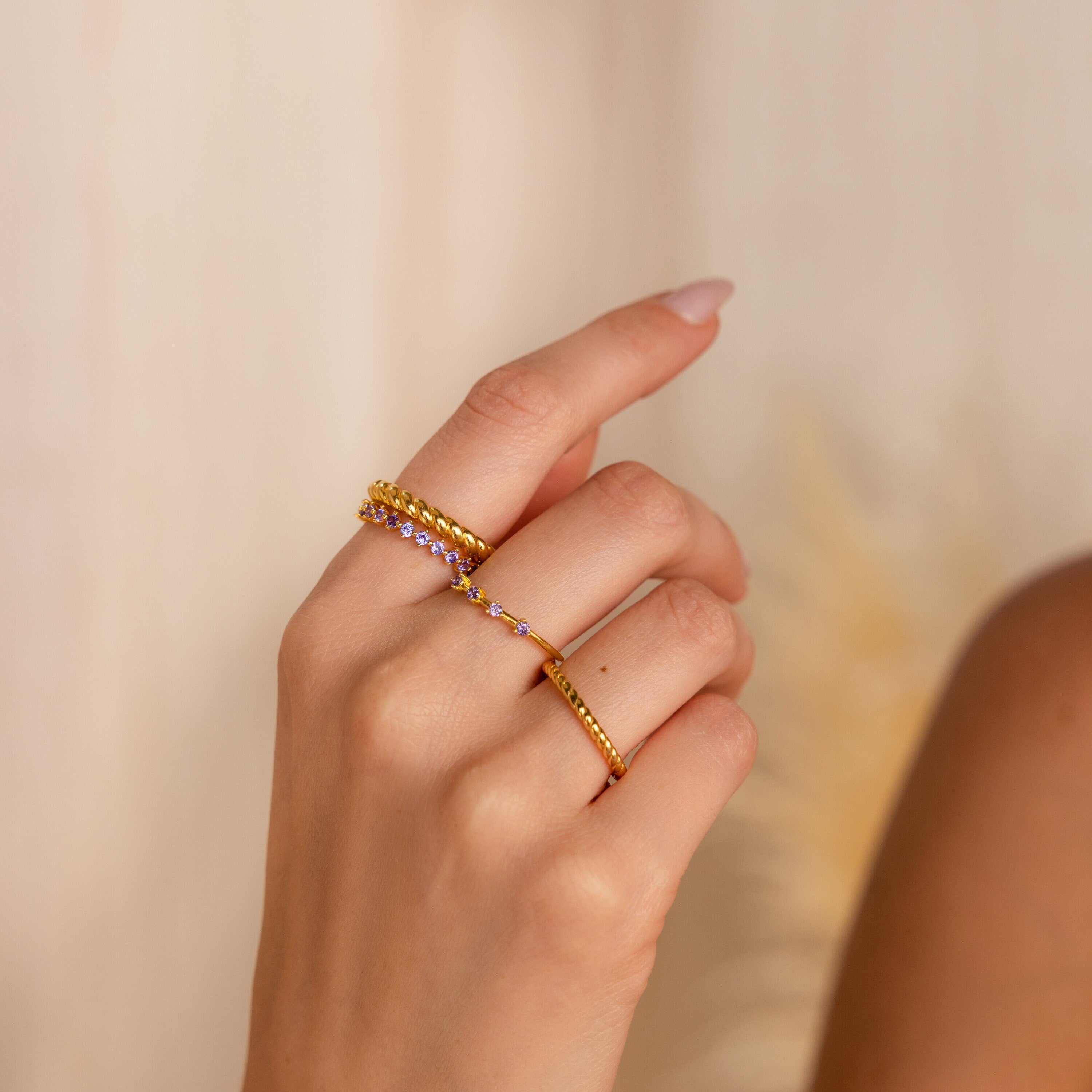A hand with three gold rings, featuring the Amethyst Eternity Ring with delicate purple stones, set against a soft beige background.