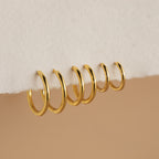Six gold Mini Bold Hoops from our collection hang on the edge of a white textured surface, showcasing their varying sizes and timeless style.