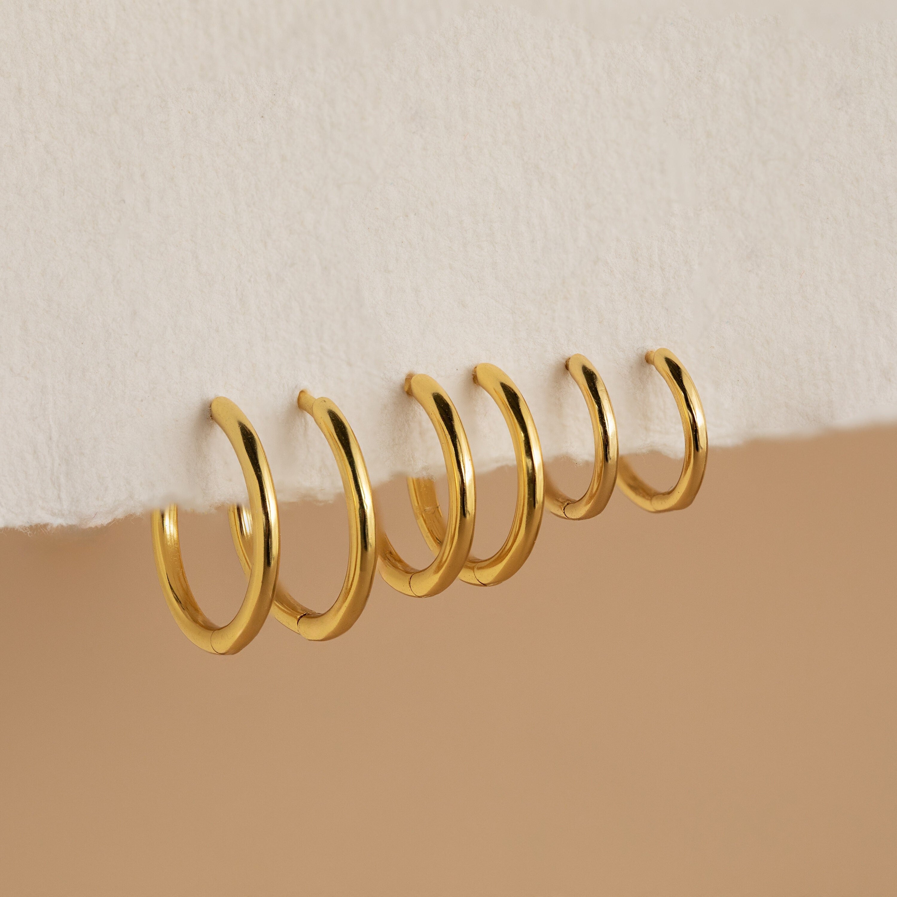 Six gold Mini Bold Hoops from our collection hang on the edge of a white textured surface, showcasing their varying sizes and timeless style.
