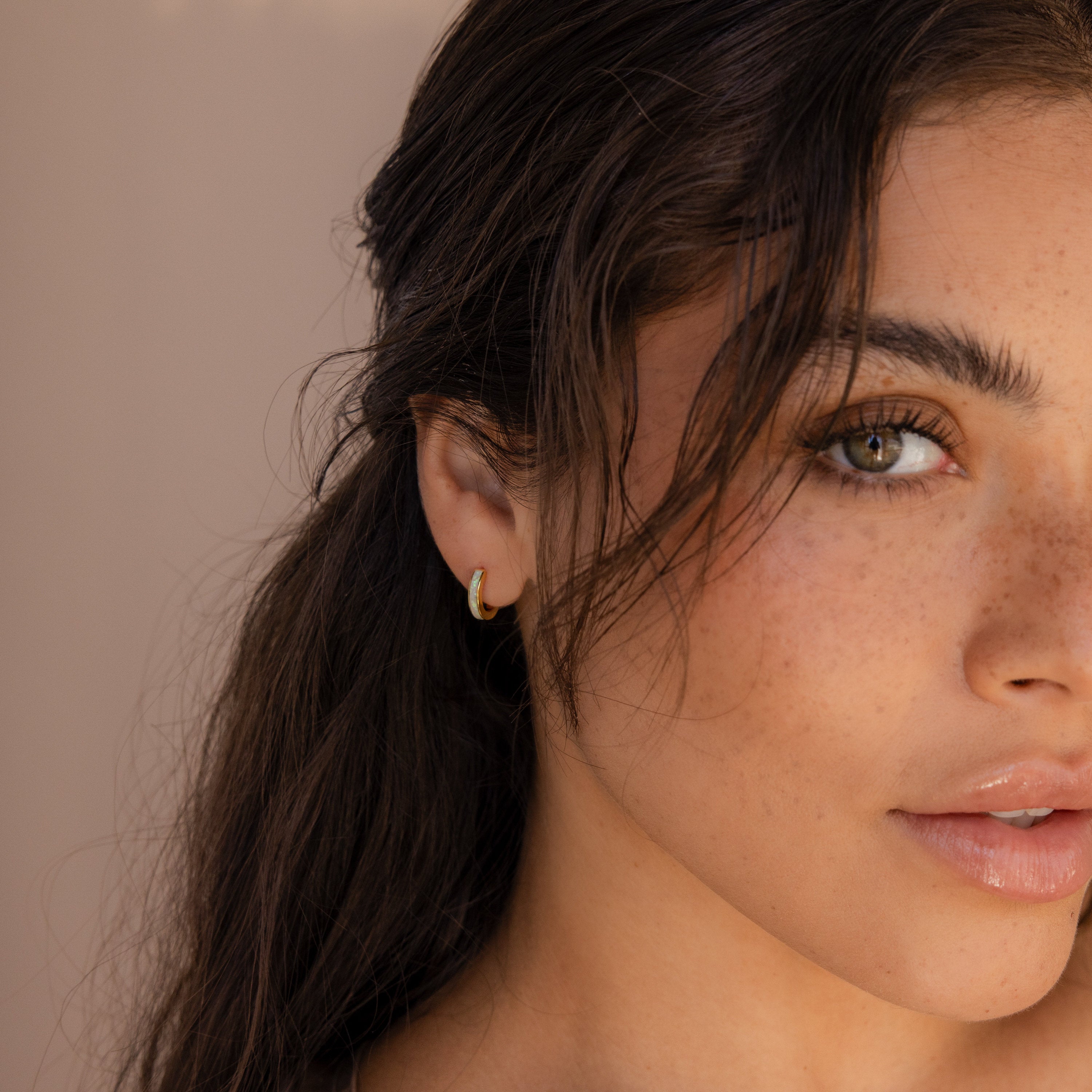 Close-up of a young woman with freckled skin, hazel eyes, and wavy brown hair wearing delicate White Opal Inlay Huggies as she looks at the camera.