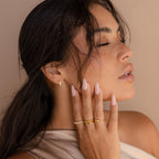 Woman with long nude nails, gold rings, and White Opal Inlay Huggies touches her face with eyes closed against a beige background.