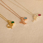 Three Initial Heart Birthstone Necklaces, each with an "M" letter pendant and a custom birthstone charm, are displayed on a textured beige background.