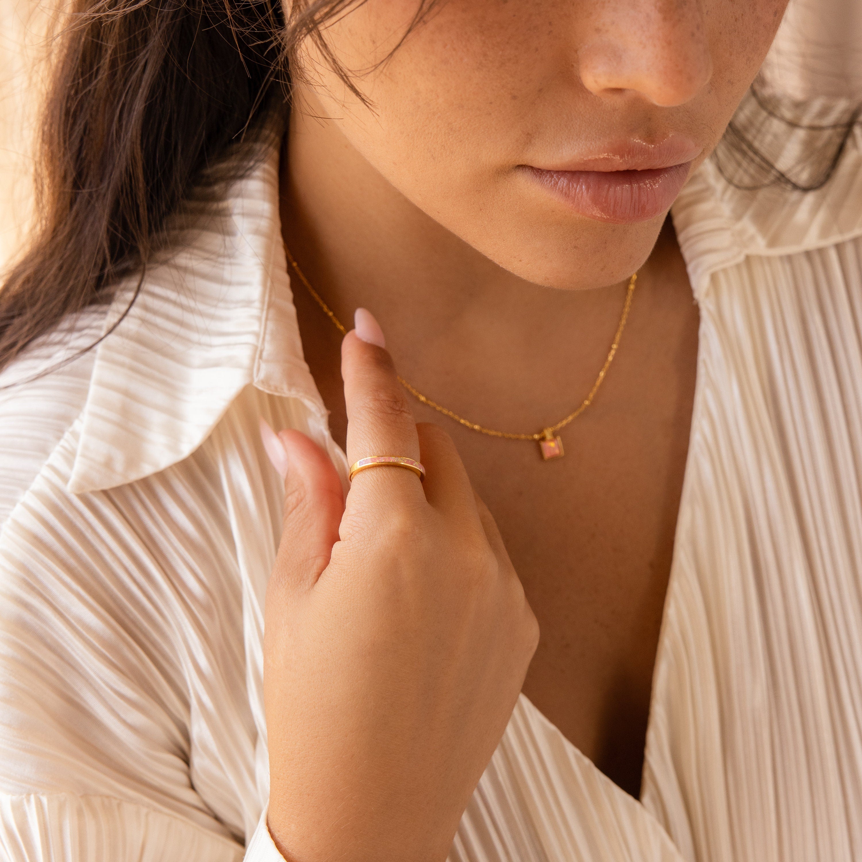 A woman in a white pleated blouse and gold necklace gently touches her collar, showcasing the delicate Pink Opal Inlay Band on her finger.