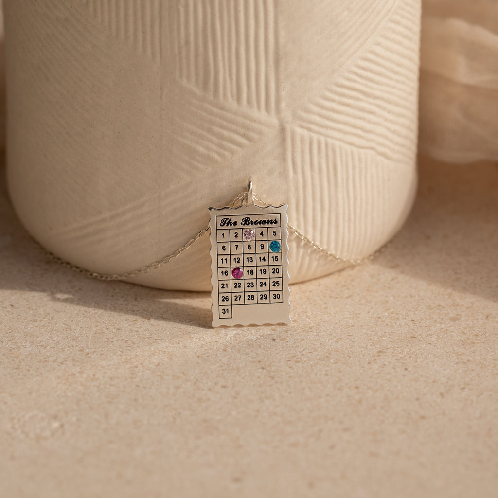 The Calendar Birthstone Necklace features a personalized calendar pendant with highlighted dates, displayed before a textured ceramic vase—a unique and meaningful gift.
