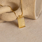 Floral Name Birthstone Necklace featuring a rectangular pendant engraved with "Hanalei," a delicate floral design, and a small gemstone—an elegant birthstone necklace shown on a stone surface.