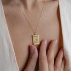 A woman wears the Calendar Birthstone Necklace on her chest, gently touching the personalized charm. She pairs it with a cream-colored top.