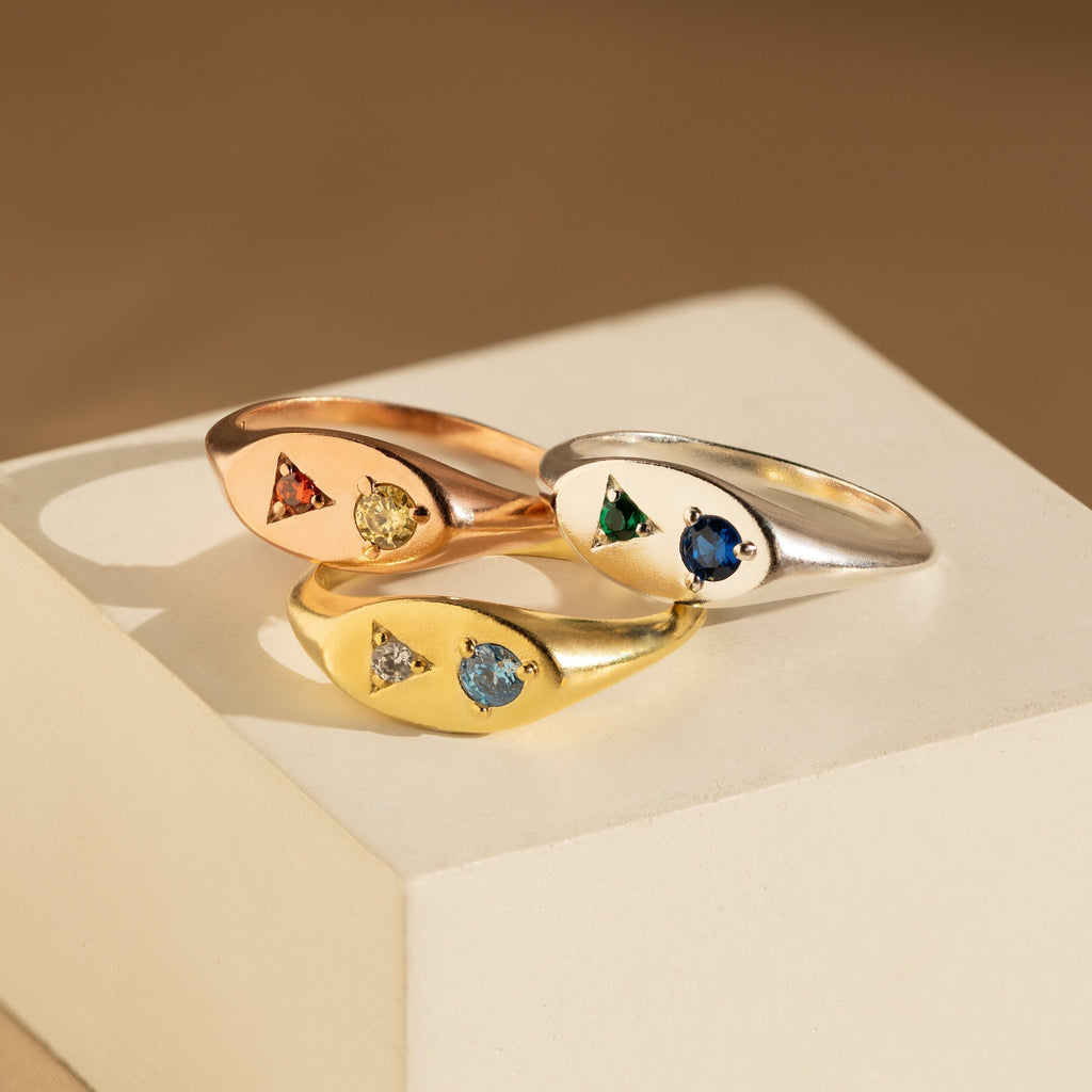 The Mixed Birthstone Signet Ring is showcased in gold, rose gold, and silver on a cream geometric block—perfect as a statement piece or for personalized jewelry styling.