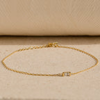 The Baguette Birthstone Bracelet features a delicate gold chain and a single rectangular clear gemstone, shown on a neutral textured surface.