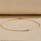 The Baguette Birthstone Bracelet features a delicate gold chain and a small rectangular blue gemstone, shown on a beige textured surface.