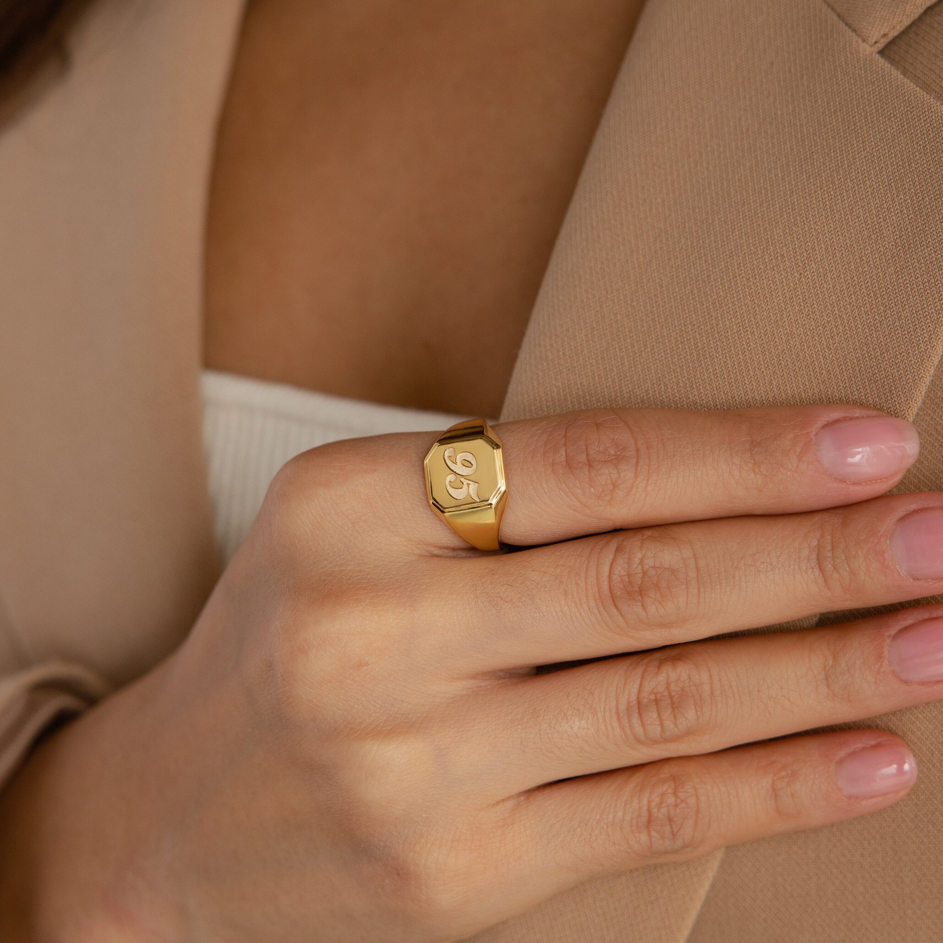 A hand with pink nails wears the Lucky Number Signet Ring, engraved with 96, touching a beige blazer—ideal statement jewelry for fans of personalized engraving.