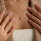 A woman wears the Initial Heart Necklace—an ideal gift for a loved one—styled with a gold heart pendant, beige blazer, and light pink manicured nails.