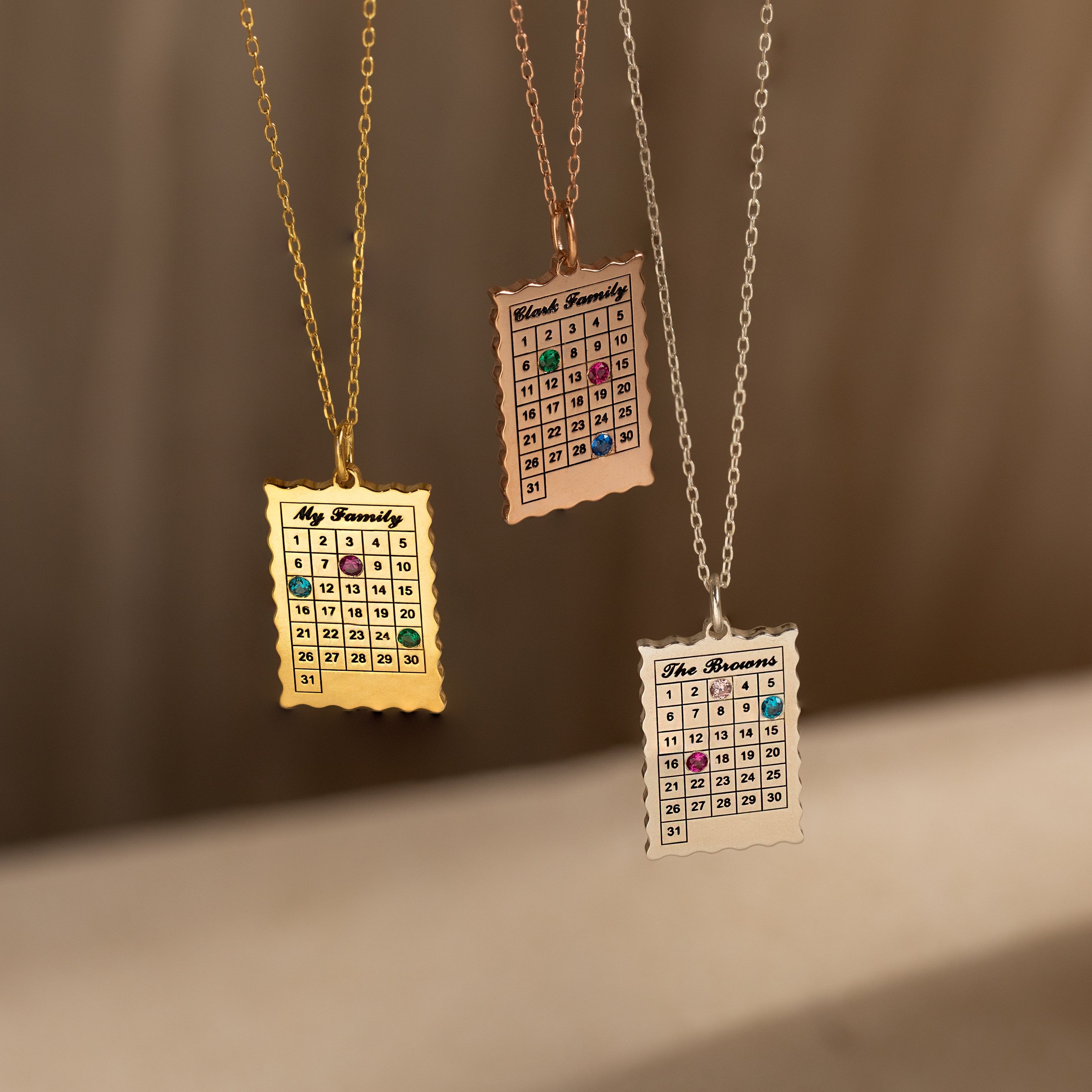 The Calendar Birthstone Necklace features gold and silver chains, each personalized with colorful birthstones and engraved family names—a unique piece of customized jewelry.
