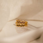 The Tiny Cluster Birthstone Rings—five gold stackable bands adorned with vibrant gemstones—are elegantly displayed on a reflective surface with beige fabric, making them perfect for anyone seeking a customizable birthstone ring.