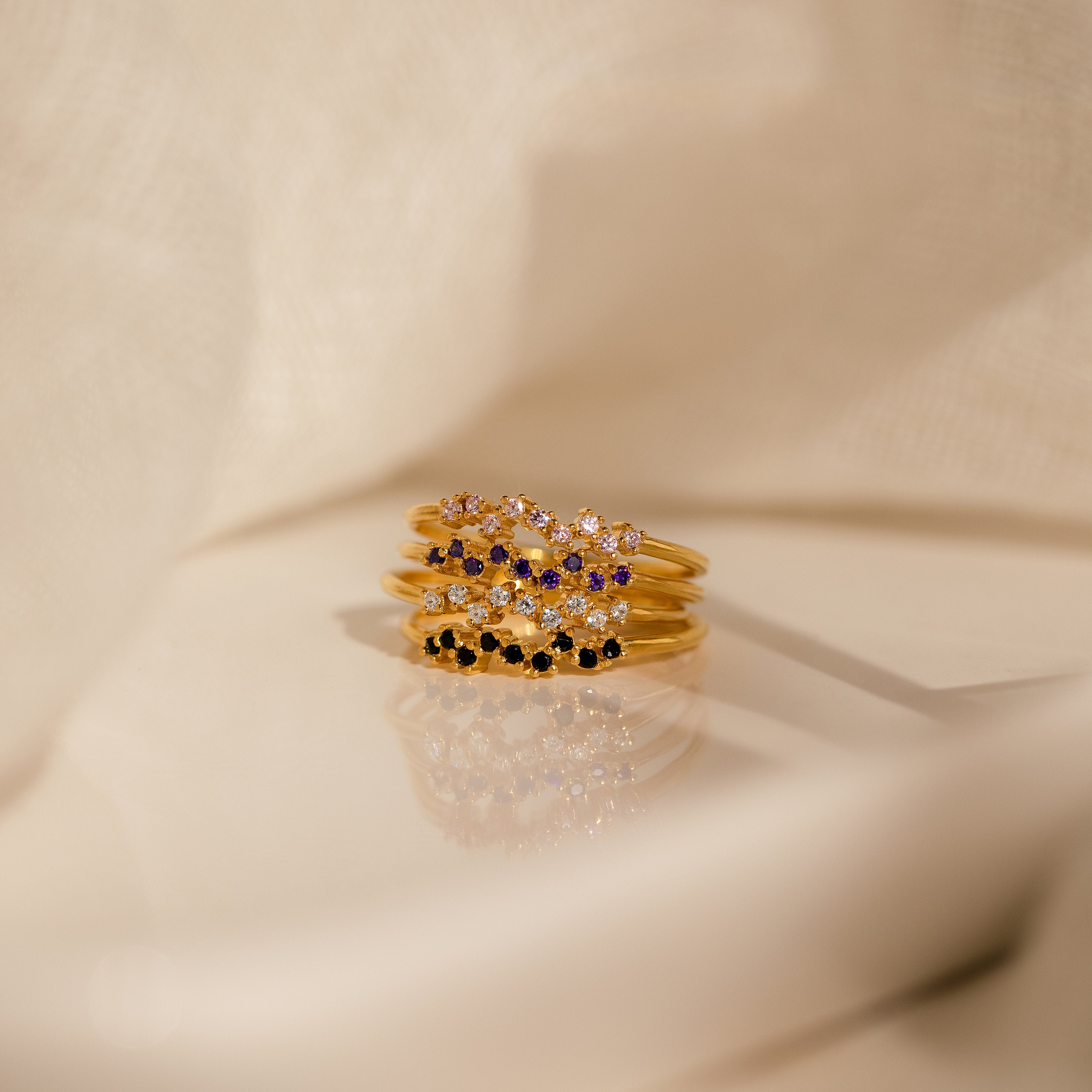 The Tiny Cluster Birthstone Rings—five gold stackable bands adorned with vibrant gemstones—are elegantly displayed on a reflective surface with beige fabric, making them perfect for anyone seeking a customizable birthstone ring.