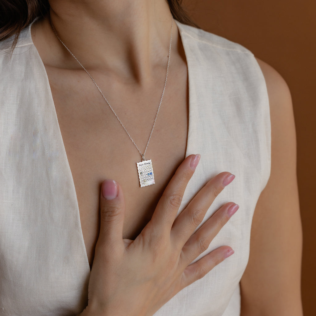 A woman in a white dress touches her chest, showcasing the Calendar Birthstone Necklace—an elegant, personalized birthstone jewelry piece.
