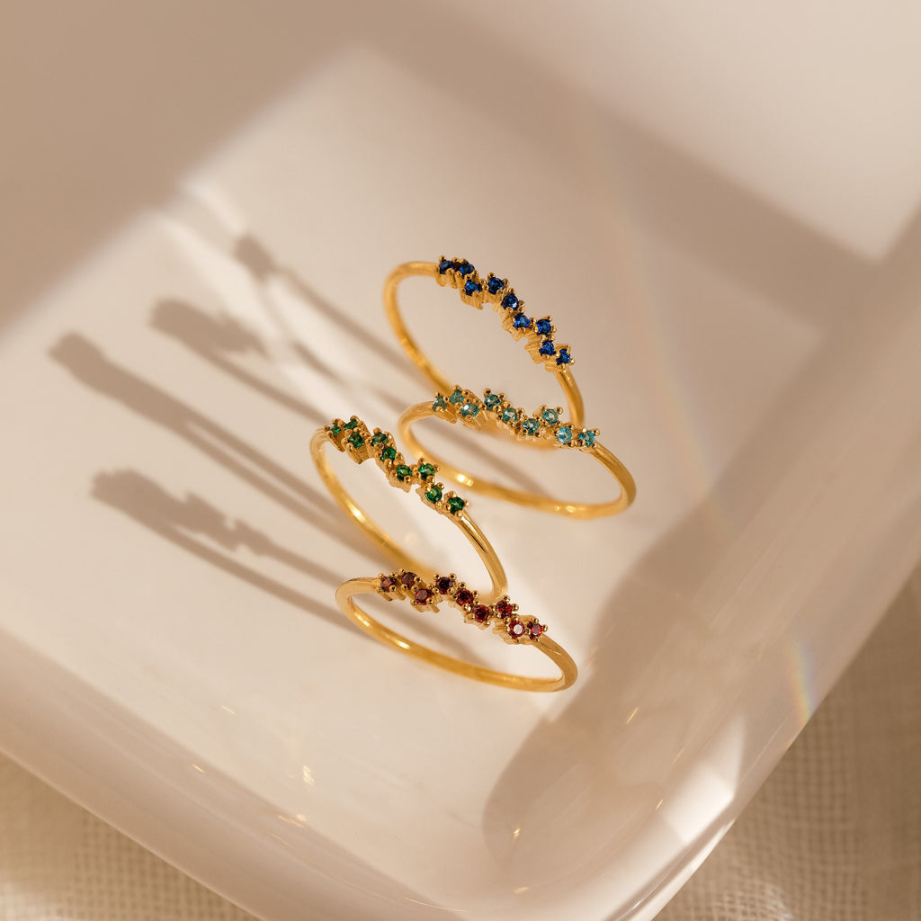 The Tiny Cluster Birthstone Ring features delicate gold with colorful gemstones, shown stacked on a white dish and casting soft shadows.
