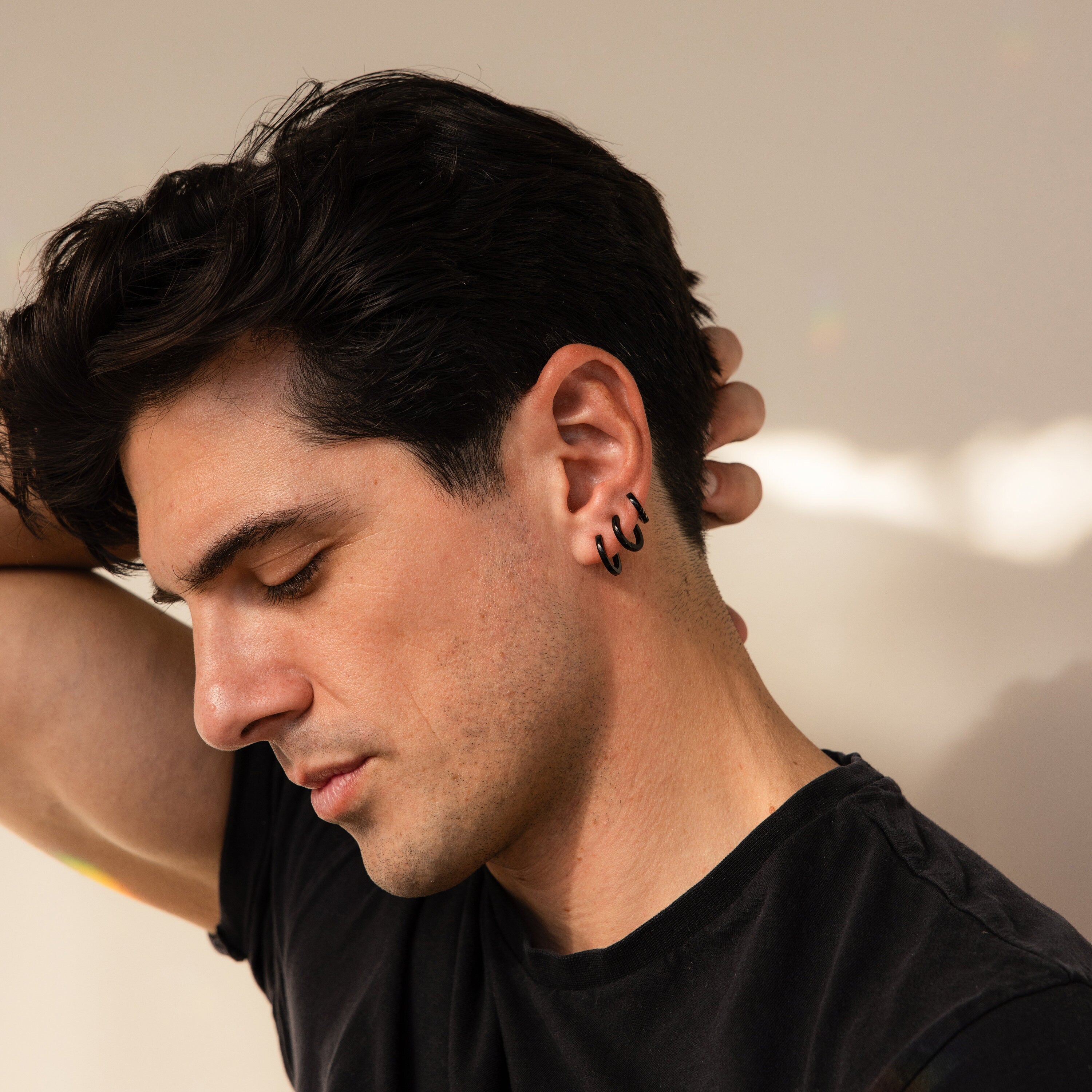A man with dark hair wears the Men's Black Hoops Set, looking down with a hand behind his head in a black t-shirt—these stylish earrings make a perfect gift for him.