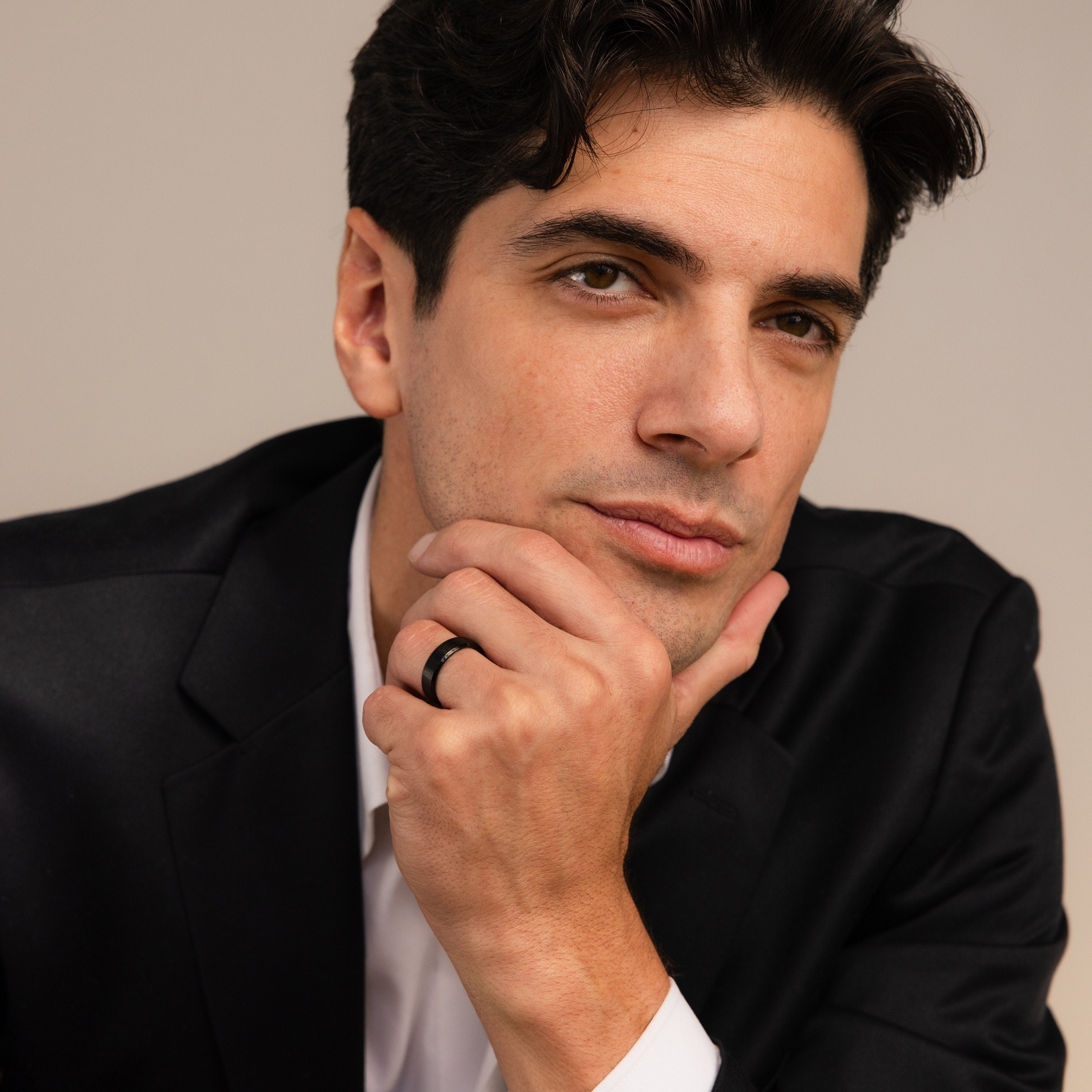 A man in a black suit, wearing the Men's Thick Black Ring, rests his chin on his hand and looks at the camera.