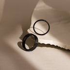 The Classic Men's Black Rings Set is displayed on a textured cream surface with soft lighting and shadows, emphasizing the sleek and minimalist design of these men's rings.