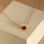 The Heart Birthstone Necklace features a vibrant red gemstone pendant, elegantly showcased on a light-colored geometric block—a perfect birthday gift or timeless personalized jewelry piece.