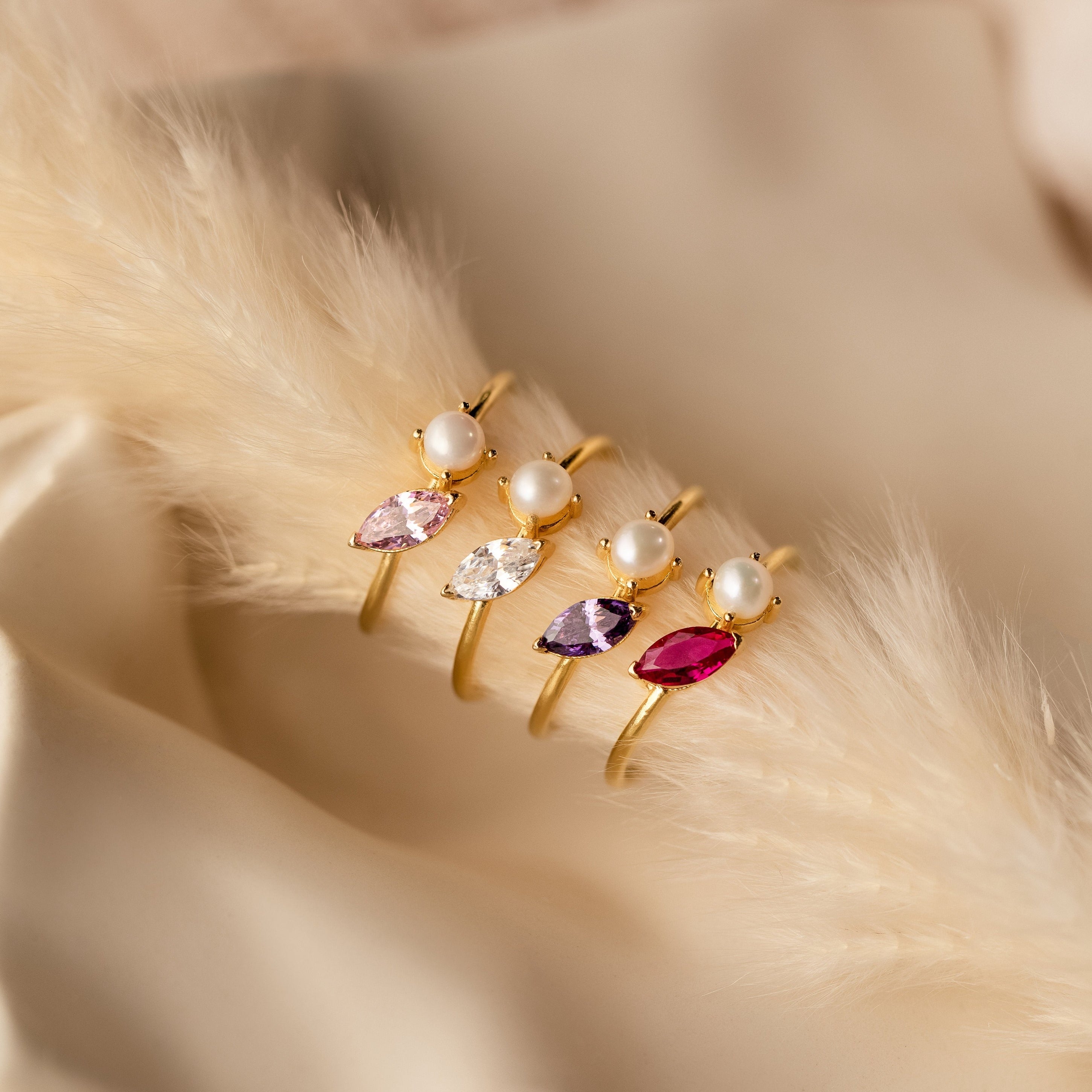 The Marquise Birthstone & Pearl Ring, featuring pearls and colored gemstones, is displayed with other gold rings on soft beige fabric and fur.