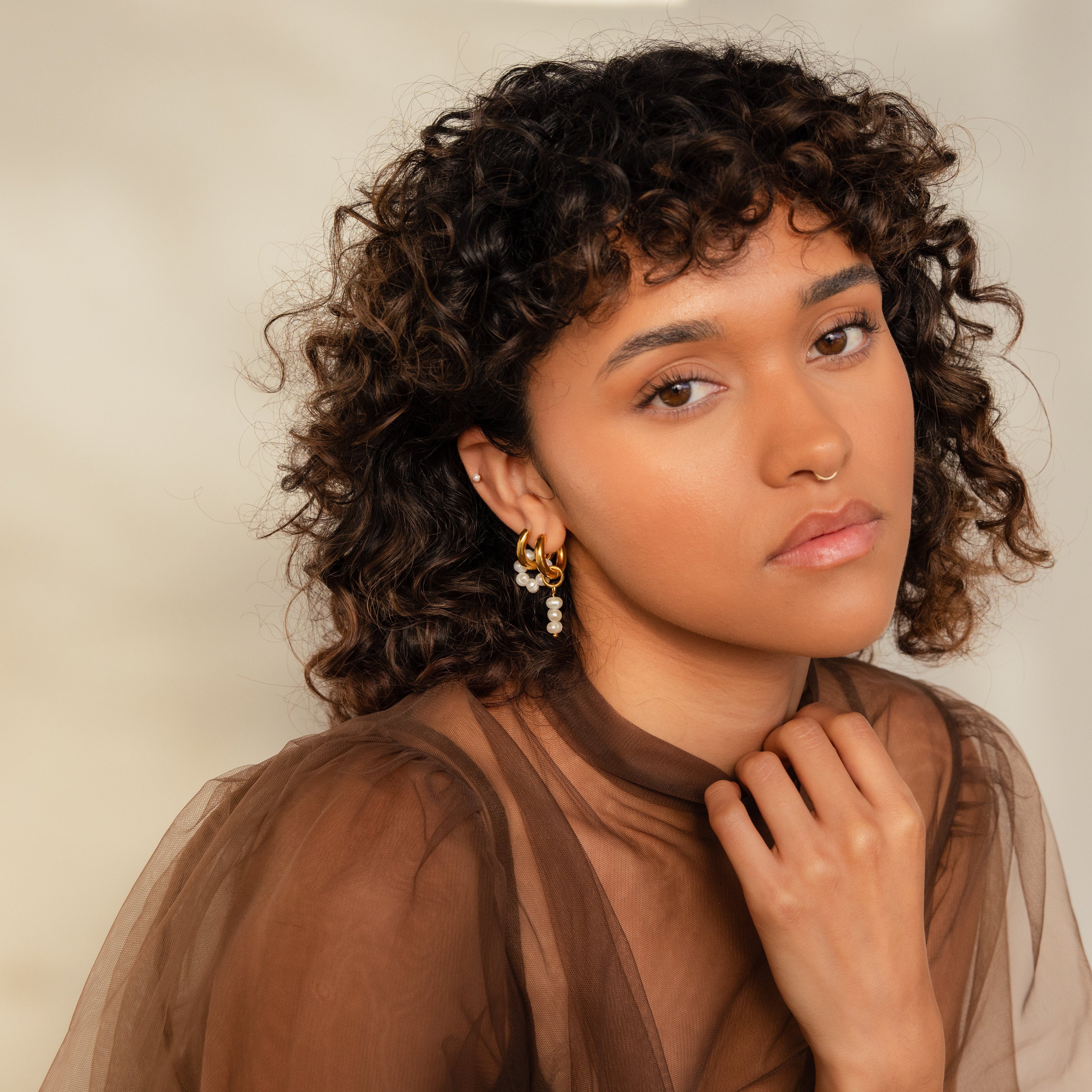 Woman with curly hair wearing Mismatched Pearl Drop Hoops and a sheer brown top, gazing thoughtfully at the camera.
