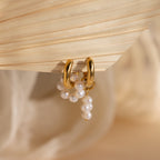 Mismatched Pearl Drop Hoops feature gold hoops with dangling clusters of white pearls, creating a striking statement look, elegantly displayed on a textured beige surface.