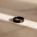 The Men's Thick Black Ring sits on a sunlit, light marble surface with soft shadows—an elegant option for men or as a modern wedding band.