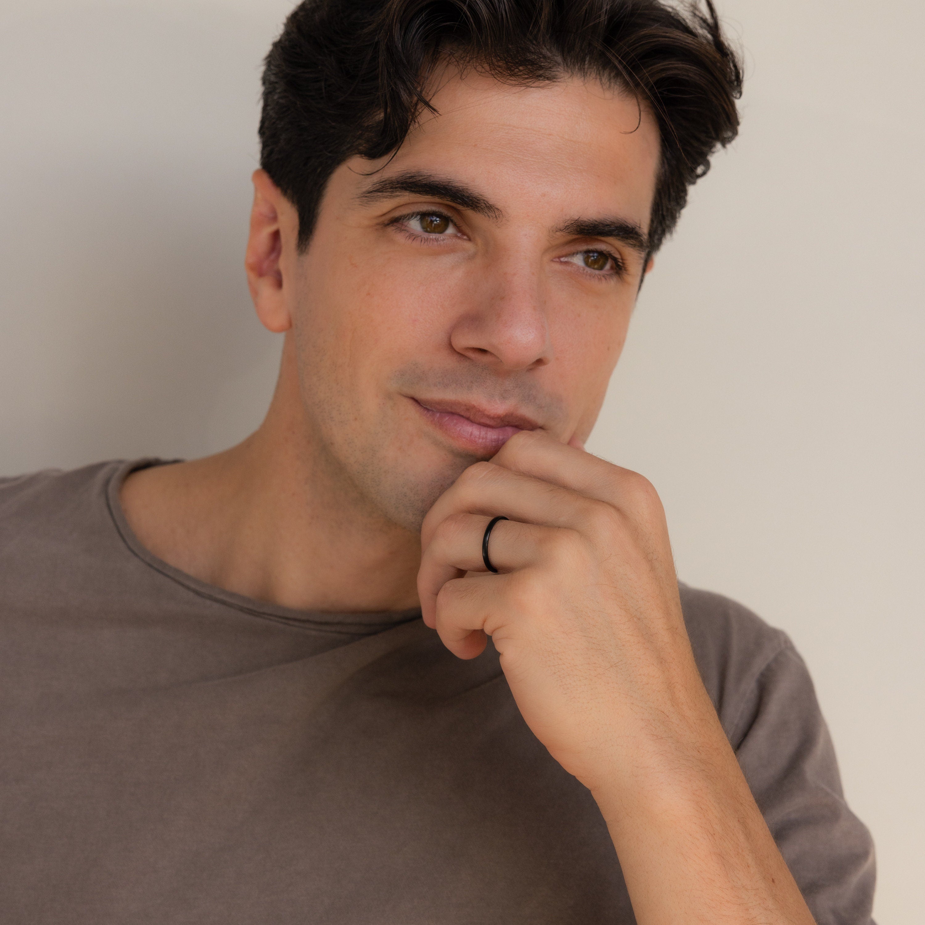 A man with dark hair in a brown shirt rests his chin on his hand, looking thoughtful—his Classic Men's Black Rings Set adds a subtle touch of style.