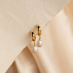 Pearl Drop Hoops feature chunky gold hoops in a minimalist style, accented with dangling white pearls, displayed on a textured beige surface.