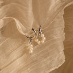 The Mismatched Pearl Drop Hoops feature two silver statement hoops adorned with small pearls, displayed on a textured beige fabric background—ideal as an elegant bridesmaid gift.