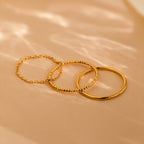 Three delicate rings from the Textured Stacking Ring Set, each featuring a unique texture, are arranged on a beige surface under soft natural lighting.