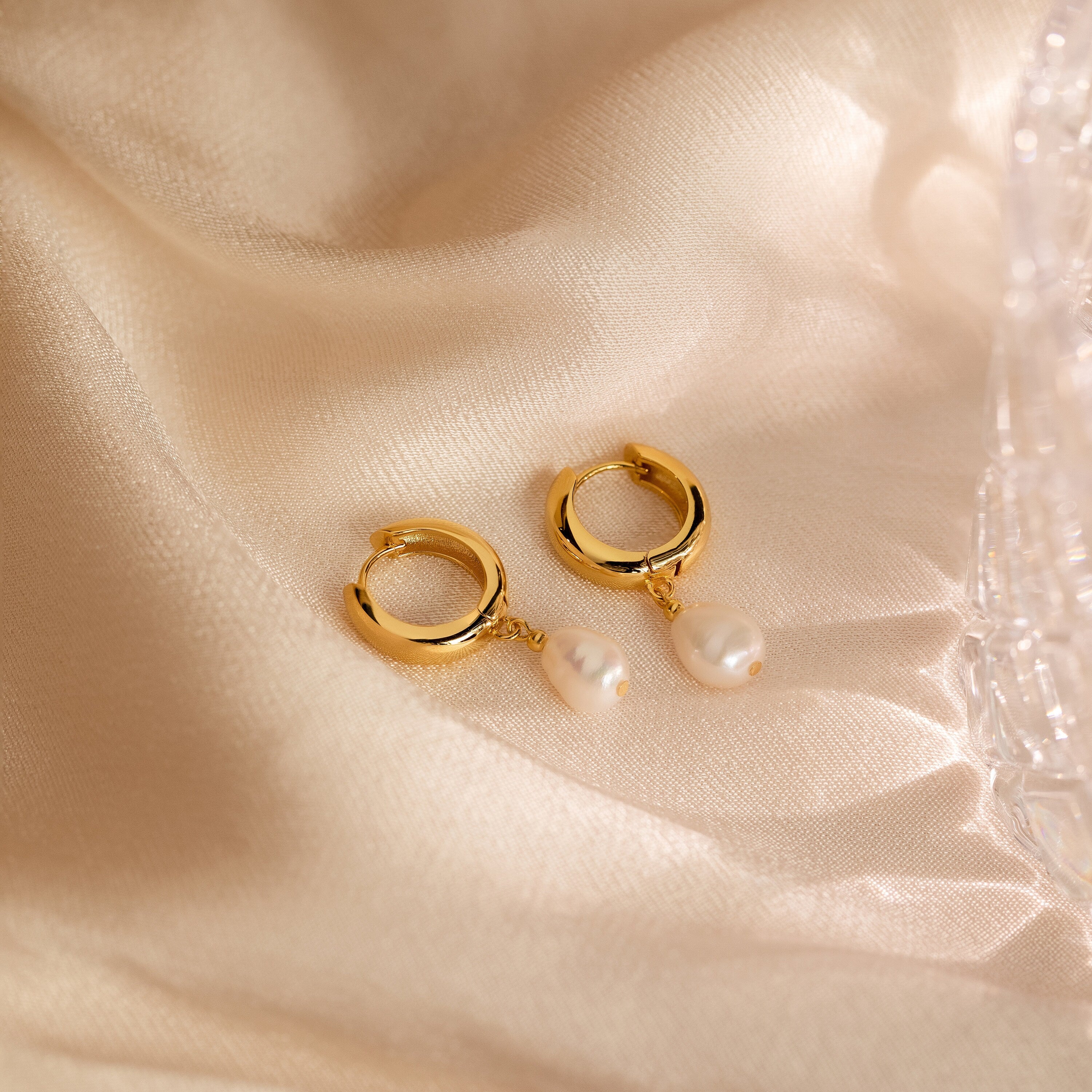 Pearl Drop Hoops with chunky design and dangling pearls rest on soft, beige satin, highlighting their minimalist drop earring style.